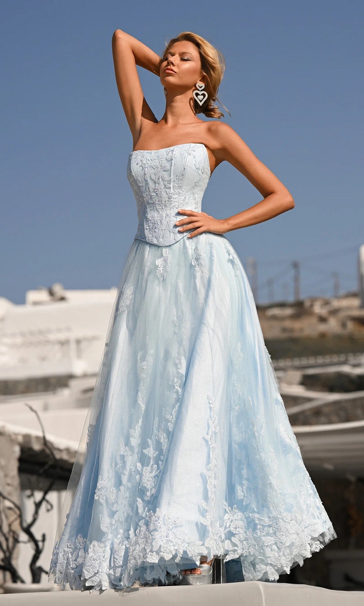 A woman poses outdoors in the Portia and Scarlett PS26061 long formal dress—a strapless, light blue A-line gown with a fitted corset bodice and full skirt—styled with large heart-shaped earrings.